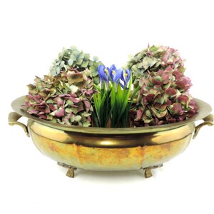 Pode incluir: Um vaso de latão com três pés e duas alças. O vaso está cheio de hortênsias secas em tons de rosa, verde e roxo, e íris azuis.