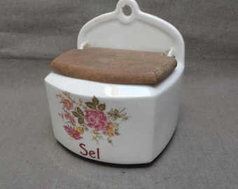 May include: White ceramic salt keeper with a wooden lid. The front of the keeper features a floral design with pink and yellow roses and the word "Sel" in red. The keeper has a curved top with a hole for hanging.