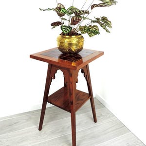 Pode incluir: Um suporte de plantas de madeira com um topo quadrado e uma prateleira inferior. O suporte tem entalhes intrincados nas pernas e é pintado de cor marrom escura. Um vaso de latão com uma planta está na prateleira superior.