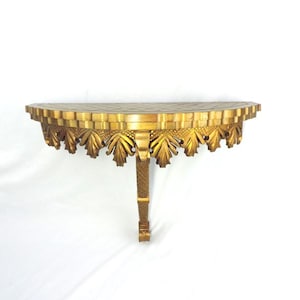 May include: A gold-coloured, semi-circular wall shelf with ornate leaf and geometric detailing. The shelf is supported by a single, textured leg. The shelf's design is reminiscent of classical architecture.