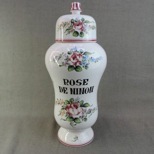 May include: A white ceramic apothecary jar with a lid, decorated with floral rose designs in red, pink, and blue. The jar has the text "ROSE DE NINON" in black. A decorative item.