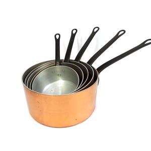 May include: A collection of five copper saucepans with black handles, arranged in a nested configuration. The copper exterior complements the silver-coloured interior. The handles are long and curved, and the pans vary in size.