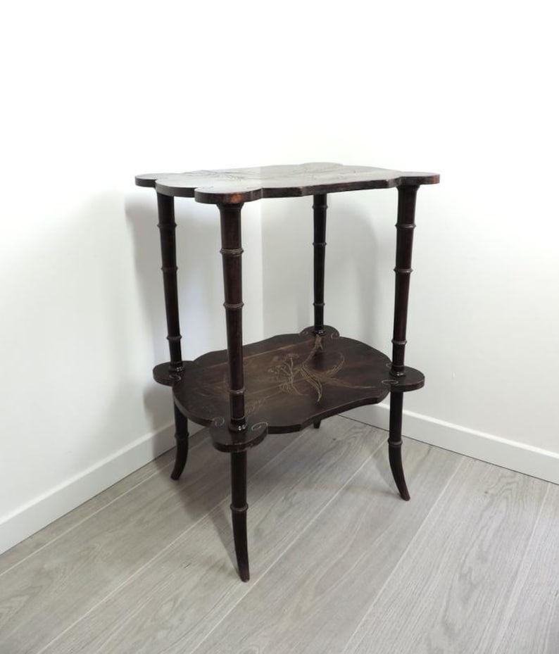 Puede incluir: Una mesa auxiliar de dos niveles en color marr&oacute;n oscuro con un dise&ntilde;o de bordes festoneados. Las cuatro patas presentan una textura segmentada estilo bamb&uacute;. Ambas superficies est&aacute;n decoradas con un delicado patr&oacute;n floral incrustado.