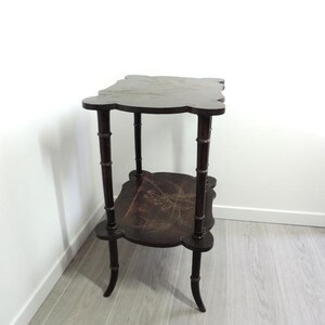 Puede incluir: Una mesa auxiliar de dos niveles en color marr&oacute;n oscuro con un dise&ntilde;o de bordes festoneados. La mesa cuenta con cuatro patas con forma de tallos de bamb&uacute; y dos estantes rectangulares, cada uno decorado con una sutil ilustraci&oacute;n floral clara.
