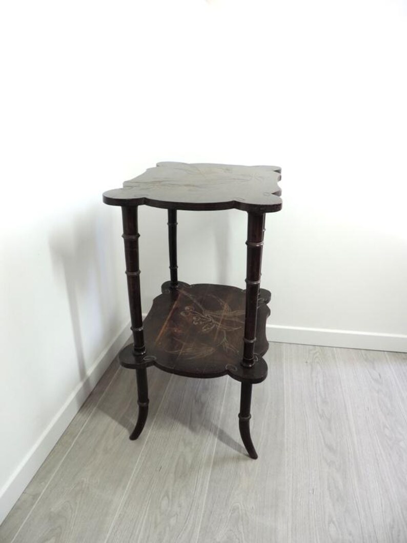 Puede incluir: Una mesa auxiliar de dos niveles en color marr&oacute;n oscuro con un dise&ntilde;o de bordes festoneados. La mesa cuenta con cuatro patas con forma de tallos de bamb&uacute; y dos estantes rectangulares, cada uno decorado con una sutil ilustraci&oacute;n floral clara.