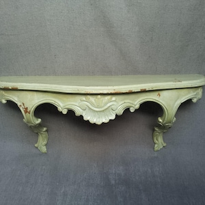 May include: A wall-mounted console shelf in a pale sage green finish with a curved top surface. The piece features ornate, carved scrollwork and shell-like detailing along the front apron, supported by two decorative, curved bracket legs.