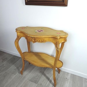May include: A vintage, light-yellow, two-tiered side table with ornate, curved legs. The top surface features hand-painted pink floral accents. The table has a lower shelf and is made of wood. The table is approximately 30 inches long.