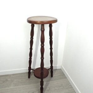 May include: A tall, dark brown wooden plant stand featuring a circular top surface and a smaller circular lower shelf. The stand is supported by three slender, turned legs with decorative spindle detailing that connect the top and bottom tiers.
