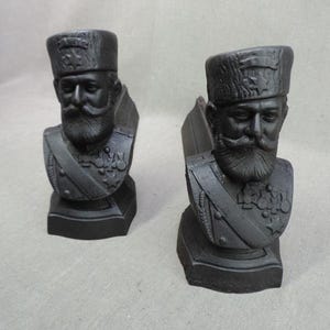 Puede incluir: Dos bustos negros de un hombre con bigote y barba, con uniforme militar y sombrero. Los bustos están sobre una base cuadrada y tienen rasgos faciales detallados y decoraciones uniformes.