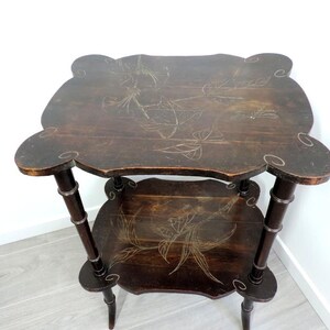Puede incluir: Una mesa auxiliar de madera de dos niveles en color marr&oacute;n oscuro con bordes festoneados. Ambas superficies presentan grabados detallados de flores y lib&eacute;lulas en tonos claros. La mesa se sostiene sobre cuatro patas delgadas y segmentadas.