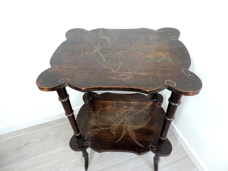 Puede incluir: Una mesa auxiliar de madera de dos niveles en color marr&oacute;n oscuro con bordes festoneados. Ambas superficies presentan grabados detallados de flores y lib&eacute;lulas en tonos claros. La mesa se sostiene sobre cuatro patas delgadas y segmentadas.