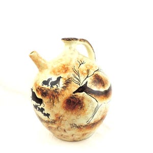May include: A ceramic jug with a brown and white glaze. The jug features a black silhouette of a deer and other animals in a cave painting style.