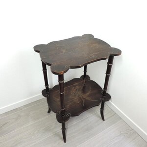 Puede incluir: Una mesa auxiliar de dos niveles en color marr&oacute;n oscuro con bordes festoneados en ambos estantes. La madera presenta motivos florales pintados a mano en tonos claros y cuatro patas con una apariencia segmentada estilo bamb&uacute;.