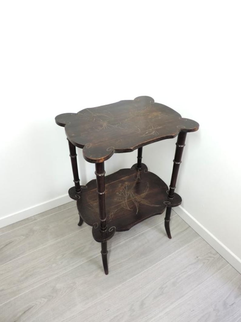 Puede incluir: Una mesa auxiliar de dos niveles en color marr&oacute;n oscuro con bordes festoneados en ambos estantes. La madera presenta motivos florales pintados a mano en tonos claros y cuatro patas con una apariencia segmentada estilo bamb&uacute;.