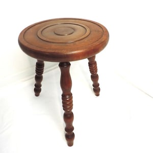 May include: A small, round wooden stool with a dark brown finish. The stool has a circular top with a carved design and three turned legs. The legs are evenly spaced and provide stability. The stool is suitable for various uses.