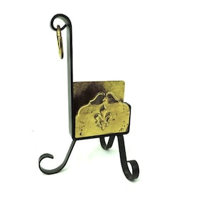 Puede incluir: Un porta llaves de metal negro con un diseño de flor de lis dorada. El soporte tiene un anillo en la parte superior para colgar llaves y un soporte con tres patas.