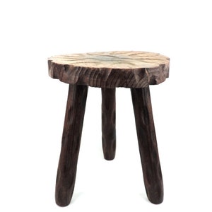 Puede incluir: Un taburete de madera rústico con una tapa redonda de madera natural y tres patas robustas. El taburete es de color marrón oscuro y tiene una superficie rugosa y texturizada.