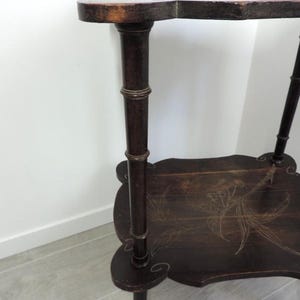 Puede incluir: Una mesa auxiliar de madera de dos niveles en color marr&oacute;n oscuro con un dise&ntilde;o de bordes festoneados. La mesa cuenta con cuatro patas con textura estilo bamb&uacute; y un delicado patr&oacute;n floral incrustado en color claro en la repisa inferior.