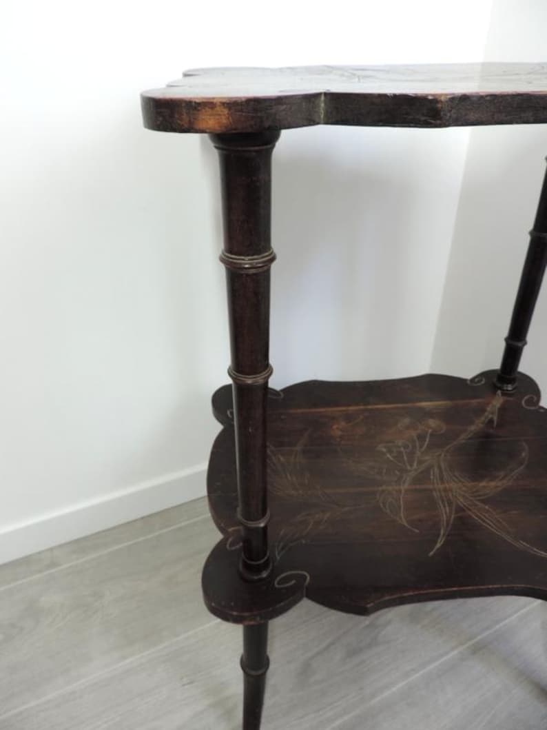 Puede incluir: Una mesa auxiliar de madera de dos niveles en color marr&oacute;n oscuro con un dise&ntilde;o de bordes festoneados. La mesa cuenta con cuatro patas con textura estilo bamb&uacute; y un delicado patr&oacute;n floral incrustado en color claro en la repisa inferior.