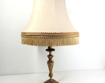 May include: A gold table lamp with a white lampshade with a gold fringe trim. The lamp base has an ornate design.