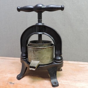 Pode incluir: Uma prensa de frutas vintage de ferro fundido preto com um mecanismo de parafuso central e uma alça horizontal. O dispositivo inclui uma cesta de metal cilíndrica com lados perfurados e um pequeno bico na base, apoiado por três pernas.