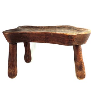 May include: A rustic wooden stool with a unique, irregular shape and four turned legs. The stool is made of dark brown wood and has a natural, weathered finish.