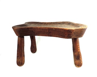 Puede incluir: Un taburete de madera rústico con una forma única e irregular y cuatro patas torneadas. El taburete está hecho de madera marrón oscura y tiene un acabado natural y desgastado.