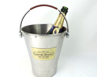 May include: A silver champagne bucket with a brown handle holds a bottle of Laurent-Perrier Champagne. The bucket has a label with the brand name and the word "Champagne". The bucket is made of metal and has a simple, elegant design.