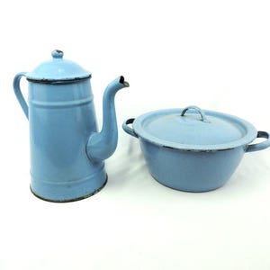 Può includere: Una caffettiera in smalto azzurro con coperchio e una ciotola abbinata con coperchio. La caffettiera ha un lungo beccuccio e un manico. La ciotola ha due manici. Entrambi gli oggetti hanno un bordo scuro.