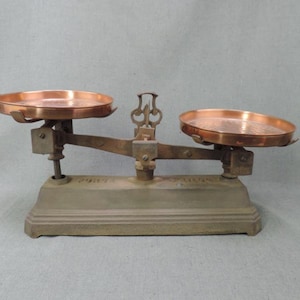 Peut inclure: Une ancienne balance avec deux plateaux en cuivre. La balance a une base grise et un bras en bronze. Les plateaux sont ronds et ont une surface légèrement texturée. La balance est conçue pour mesurer le poids.