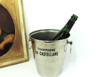May include: Silver champagne bucket with a green bottle of champagne inside. The bucket has the text "Champagne de Castellane" printed on the side.