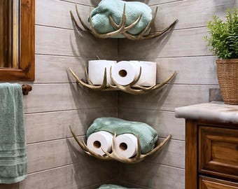 May include: Bathroom corner shelves made from deer antlers. Each shelf holds folded light green towels or rolls of toilet paper. The shelves are mounted on a light gray, wood-paneled wall, creating a rustic, lodge-style aesthetic.