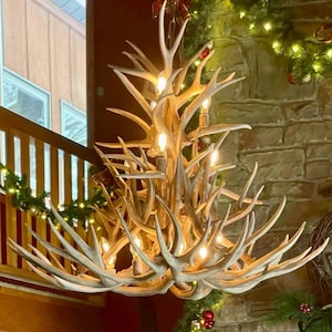 May include: A large, antler chandelier with multiple light bulbs. The chandelier is made of natural-colored antlers and hangs from a chain. The background includes a stone wall and Christmas decorations.