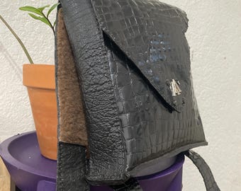 May include: A black leather backpack featuring a glossy crocodile-embossed texture on the front flap and straps, contrasted by a matte pebbled leather side panel. The bag includes a top handle, a silver-toned metal logo emblem, and a brown interior lining.