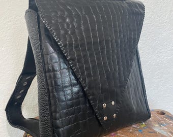 May include: A black backpack featuring a textured crocodile-embossed pattern on the front flap and a pebbled leather texture on the side panels. The bag includes a top handle, a triangular flap closure, and four silver-toned metal rivets.