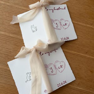 May include: Two square white cards with deckled edges, each tied with a beige chiffon ribbon. One card features a cat illustration and the text "to my husband," the other a mouse and "to my wife," both with embroidered hearts, initials, and the date 22.6.26.