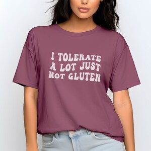 May include: A maroon t-shirt with the text "I TOLERATE A LOT JUST NOT GLUTEN" in white, stylized font. The shirt is a solid color and has short sleeves.