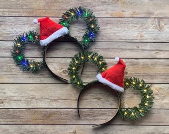 May include: Two Christmas-themed headbands with a festive design. Each headband features a black band, a wreath-shaped design with green faux pine needles, and a small Santa hat. The wreaths are adorned with small, colorful lights.