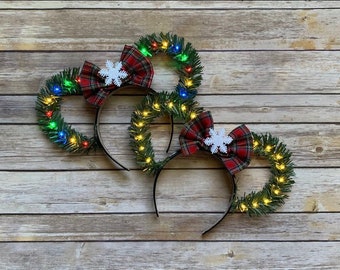 May include: Three Christmas-themed headbands with a wreath design. Each headband features a black band, a green wreath with small, colorful lights, a red plaid bow, and a white snowflake accent. The headbands are arranged on a wooden surface.