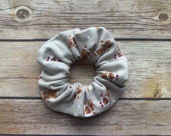 May include: A beige scrunchie with a gingerbread man pattern. The scrunchie is made of soft fabric and has a gathered look.
