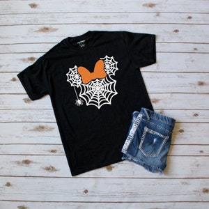 May include: Black t-shirt with a white spiderweb design in the shape of Mickey Mouse ears. The bow is orange.