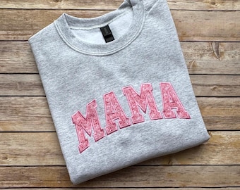 Op de afbeelding: Een grijze sweatshirt met het roze geborduurde woord "MAMA" over de borst. De sweatshirt is op een houten oppervlak gevouwen. De letters hebben een bloemenmotief.