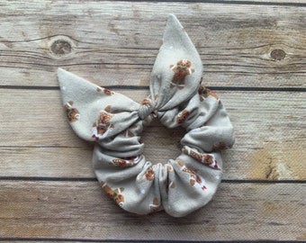 May include: A beige scrunchie with a gingerbread man pattern. The scrunchie is tied in a bow.