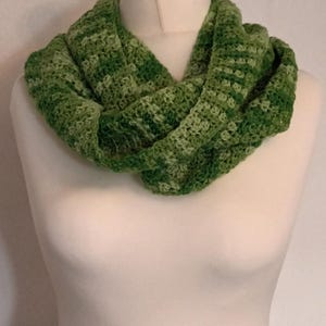 May include: A hand-crocheted infinity scarf featuring a variegated pattern of multiple shades of green, including forest, lime, and olive tones. The scarf is constructed with a textured, repeating stitch pattern and is draped around a mannequin neck.