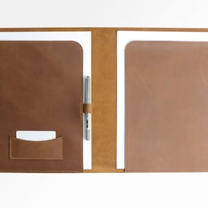 Könnte beinhalten: Ein brauner Lederordner mit einem Stifthalter und einem Kartenfach. Der Ordner ist geöffnet und enthält zwei weiße Blätter Papier.