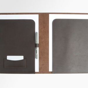 May include: A brown leather portfolio with a pen holder and a small card pocket. The portfolio is open, revealing white paper and a silver pen. The interior is a lighter brown color.