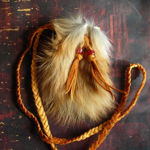 Peut inclure: Une petite pochette en fourrure avec une lanière en cuir tressé. La pochette est faite de fourrure beige et marron, avec des perles rouges et des glands en cuir beige. La lanière tressée est de couleur marron clair. Le fond est une surface sombre et texturée.