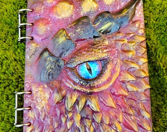 May include: A fantasy-themed journal with a dragon eye design. The cover features a textured, multi-colored design with a large, detailed dragon eye in shades of blue, gold, and purple. The journal is bound with metal rings.