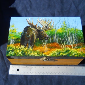 May include: A wooden box with a painting of a moose in a forest scene. The moose is brown with large antlers. The forest has green, yellow, and orange foliage. The box has a metal clasp and is approximately 12 inches long.