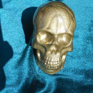 May include: A gold-colored skull figurine with detailed facial features. The skull is made of a solid material and has a metallic sheen. The skull is resting on a teal fabric.
